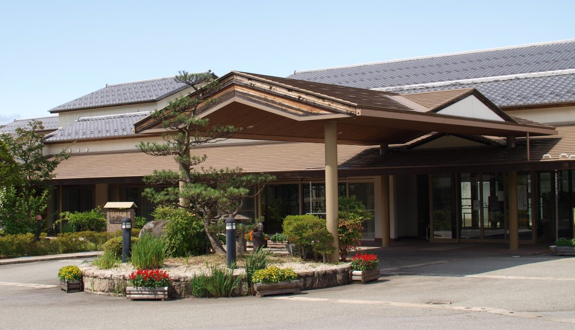 木造平屋建ての介護施設入口。庭園風の花壇と植栽が配された車寄せ